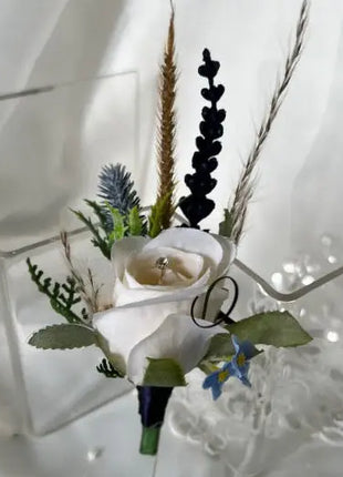 A white rose boutonniere-style floral accessory with green leaves, dried grasses, and small blue flowers, displayed on a clear acrylic stand against a white fabric backdrop.