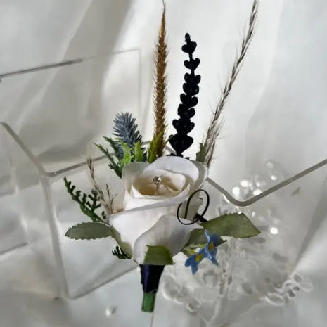 A white rose boutonniere-style floral accessory with green leaves, dried grasses, and small blue flowers, displayed on a clear acrylic stand against a white fabric backdrop.
