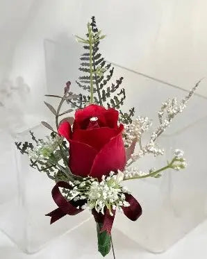 A red rose wrist corsage with white baby's breath and green foliage, tied with a dark red ribbon, shown on a clear display stand.