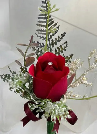 A small floral arrangement featuring a single deep red rose at the center, surrounded by green and white filler foliage, with a dark red ribbon tied at the bottom and a small clear gemstone in the rose.
