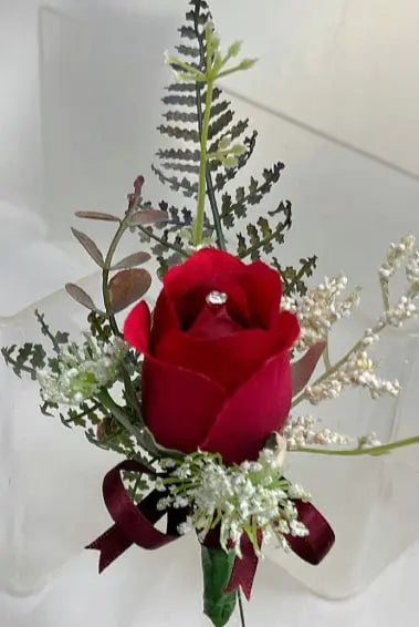 A small floral arrangement featuring a single deep red rose at the center, surrounded by green and white filler foliage, with a dark red ribbon tied at the bottom and a small clear gemstone in the rose.