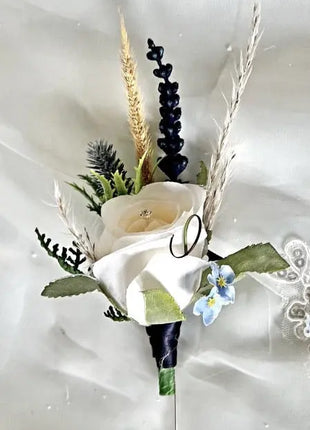 A formal boutonnier wrist flower featuring a white rose, dried wheat stalks, green foliage, and a small blue forget-me-not, pinned against a white satin fabric background.