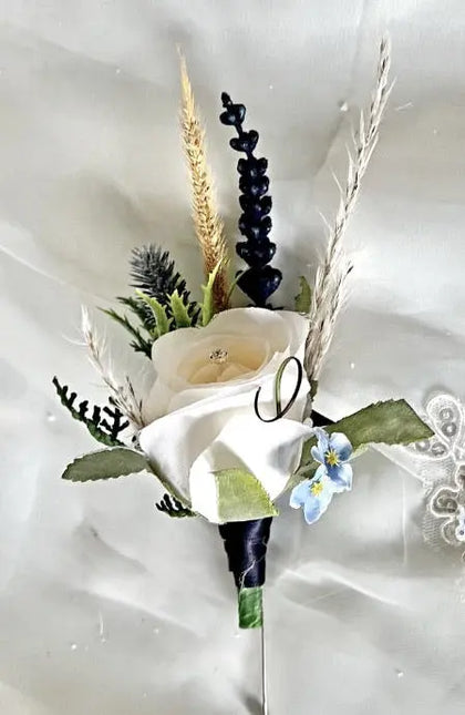 A formal boutonnier wrist flower featuring a white rose, dried wheat stalks, green foliage, and a small blue forget-me-not, pinned against a white satin fabric background.