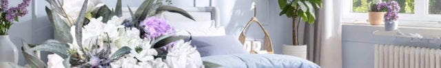 Neatly made bed with light blue bedding in a bright bedroom with a vase of flowers on a side table.