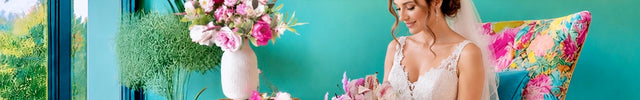 Woman in a wedding dress holding bouquet of pink  flowers in a floral room with a decorative painting of two love birds.