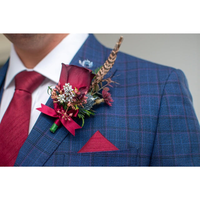 A close-up of a man's chest wearing a blue plaid suit jacket, white dress shirt, and red tie, with a matching red pocket square and a red floral boutonniere on the lapel.