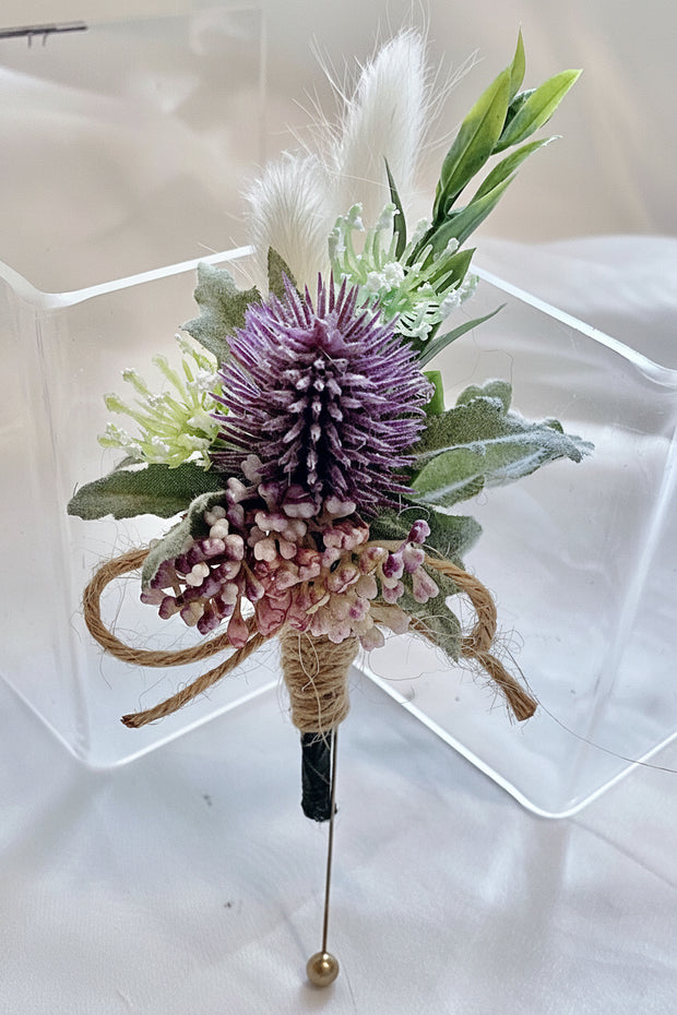 Floral boutonniere with purple thistle and greenery on a clear stand.