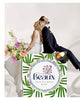 A bride and groom sit back-to-back on a white draped surface, with a large decorative sign in front that reads 'Beaux Fine Flowers Shop' and features green palm leaves. The bride wears a white gown and the groom wears a dark suit, and flowers are arranged on the left side-Beaux Faux Flowers Shop
